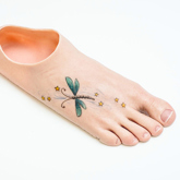 A silicone prosthetic foot with a white skin tone and a green winged dragonfly tattoo with gold stars